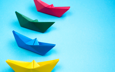 Vor einem hellblauen Hintergrund ist eine Reihe bunter Origami-Bootchen zu sehen.