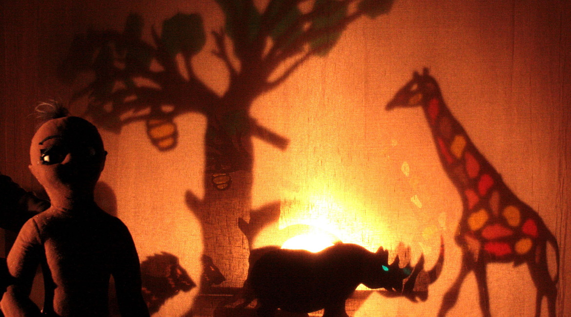  Schattentheaterszene mit Savannentierfiguren, darunter ein Nashorn, eine Giraffe und eine humanoide Figur, beleuchtet von orangefarbenem Licht.